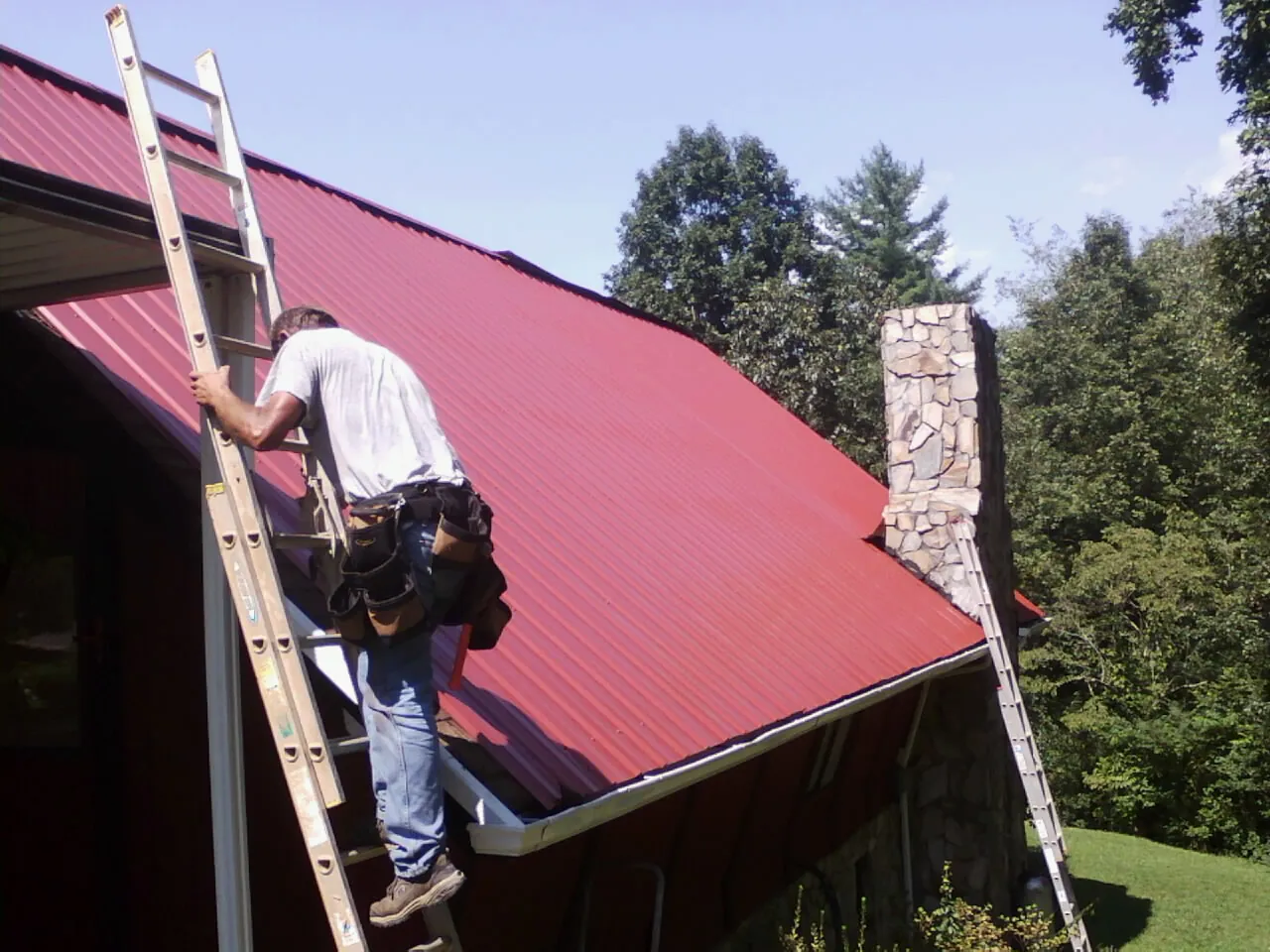 Roofer on a ladder beside a red metal roof and stone chimney — Soffit & Fascia Repair by Roofing Brothers in Mckinney